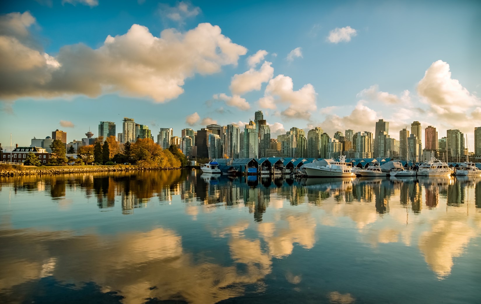 A waterfront city skyline of tall buildings and moored boats reflected in calm water under a blue sky with fluffy clouds